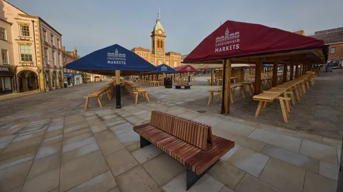 This urban landscape photograph captures Chesterfield Market Square in Derbyshire, England, during an early autumn morning. The image shows the distinctive Chesterfield Market Hall, a prominent landmark with its clock tower, situated centrally in the square. The market is set up with wooden stalls and benches, some of which are covered by canopies featuring the words "Chesterfield Markets." The photograph highlights the quietness of the square at sunrise, with soft sunlight casting long shadows across the stone paving and enhancing the architectural features of nearby historic buildings. This scene in Chesterfield, United Kingdom, focuses on the market area before the bustle of daily activity begins.
